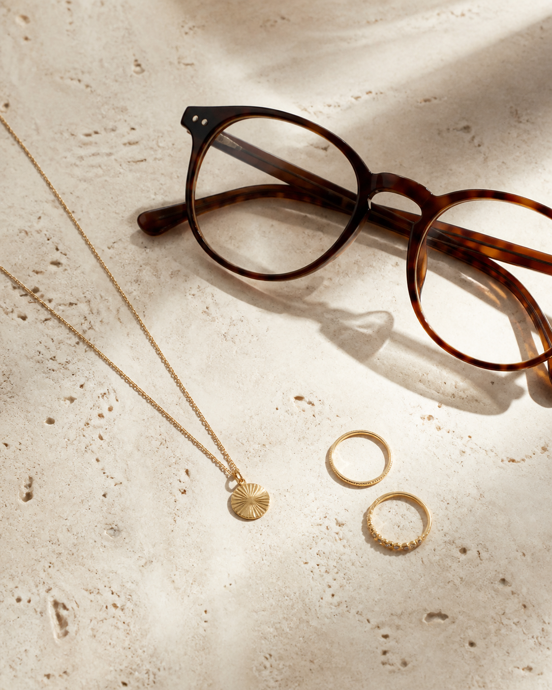 Lentes color carey con collar dorado fino y anillos delicados sobre piedra clara